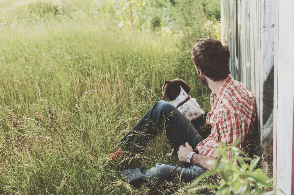 A man in plaid shirt sits with his dog in a grassy rural field, enjoying a peaceful day outdoors.